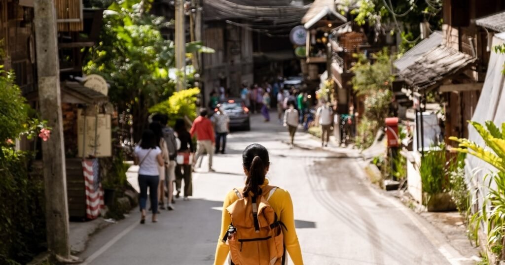 คนที่มีกระเป๋าเป้สีเหลืองเดินไปตามถนนที่พลุกพล่านซึ่งมีอาคารและต้นไม้เรียงรายอยู่สองข้างทาง โดยมีผู้คนหลายคนเดินอยู่ข้างหน้า. เที่ยวหน้าฝนโรแมนติก 