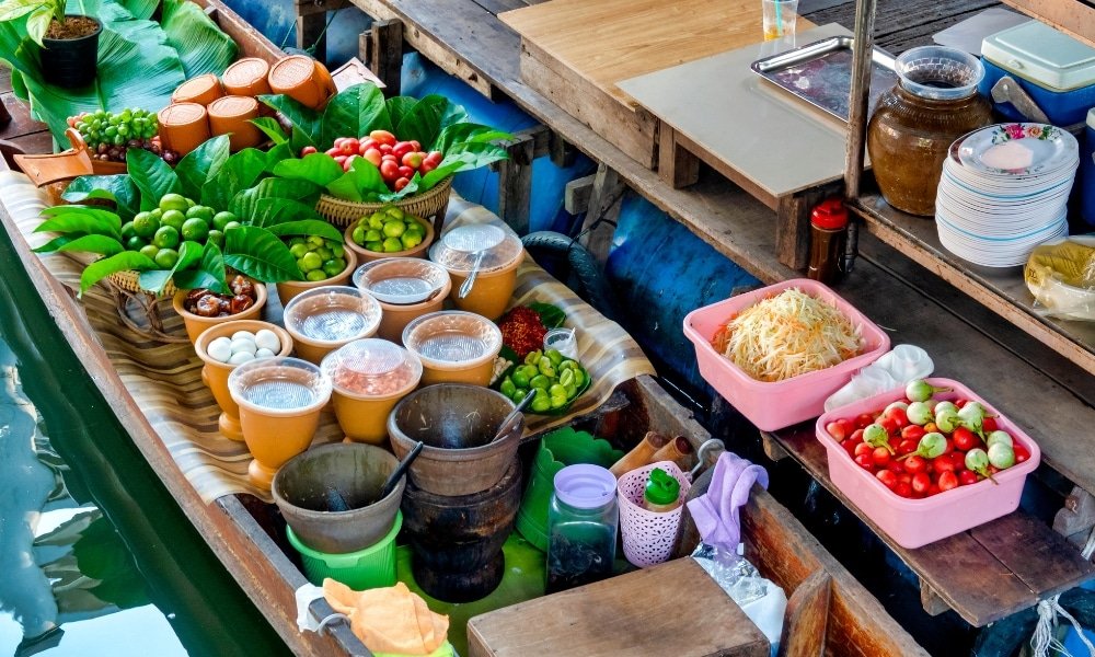 ที่ ตลาดน้ำใกล้กรุงเทพ เรือไม้ลำหนึ่งจัดแสดงผลไม้สด ผัก กระปุก และชามของส่วนผสมต่างๆ วางเคียงข้างกับเครื่องใช้และจานทั้งหมด พร้อมสำหรับการเตรียมอาหารที่มีสีสันที่ตลาดน้ำ.