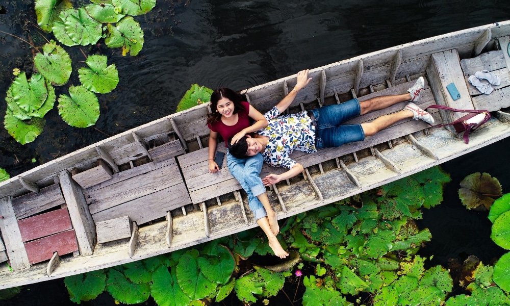 ผู้หญิงและผู้ชายนอนผ่อนคลายอยู่ในเรือไม้ ล้อมรอบด้วยดอกบัวบนผิวน้ำสีเข้ม มองจากมุมสูง สร้างความรู้สึกสงบและเสน่ห์ของ ตลาดน้ำใกล้กรุงเทพ