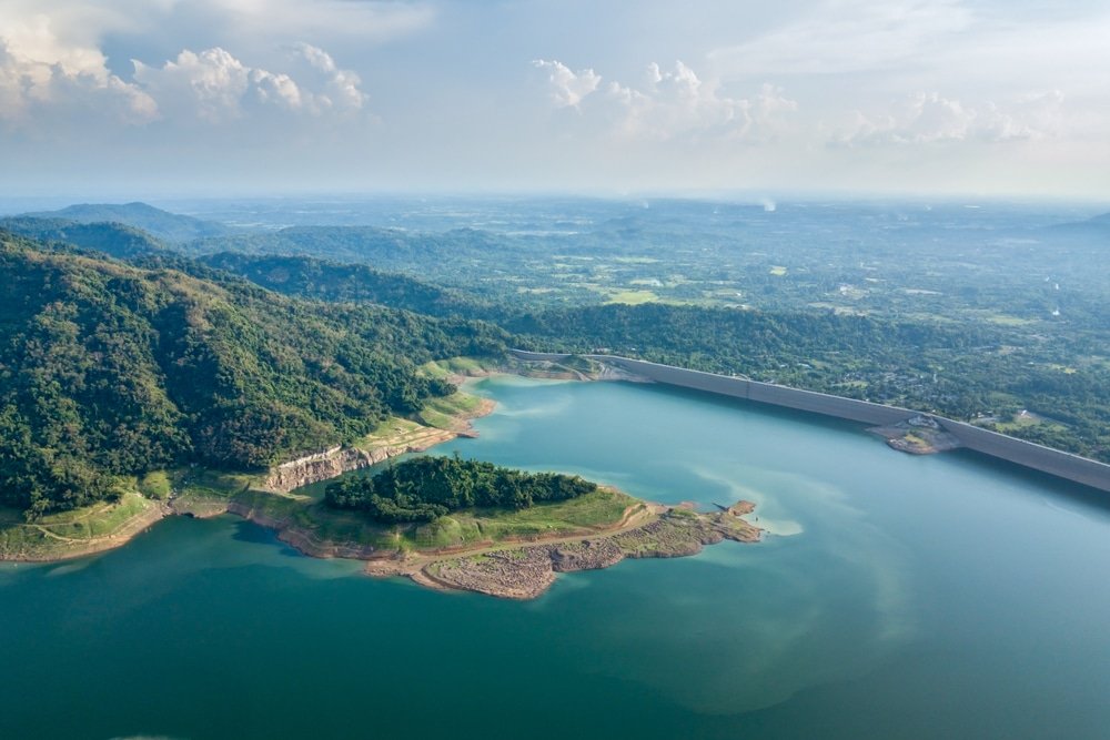 ภาพมุมสูงของอ่างเก็บน้ำขนาดใหญ่ สถานที่ท่องเที่ยวยอดนิยมทางธรรมชาติ ล้อมรอบด้วยเนินเขาที่ ที่เที่ยวธรรมชาติ ปกคลุมไปด้วยป่าและเขื่อน มีภูมิทัศน์กว้างใหญ่และท้องฟ้าที่มีเมฆปกคลุมเป็นฉากหลัง.