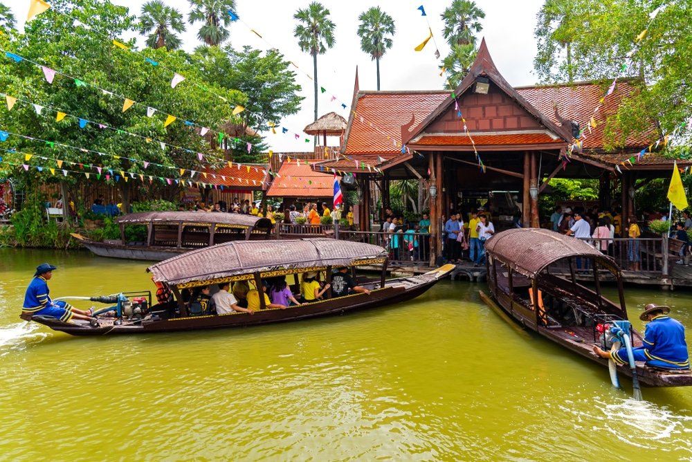 เรือไม้ที่มีผู้โดยสารแล่นผ่านคลองใกล้กับอาคารสไตล์ไทยดั้งเดิมที่ ตลาดน้ำใกล้กรุงเทพ ที่คึกคัก ผู้คนมารวมตัวกันบนท่าเรือและธงสีสันสดใสถูกแขวนไว้เหนือศีรษะ.