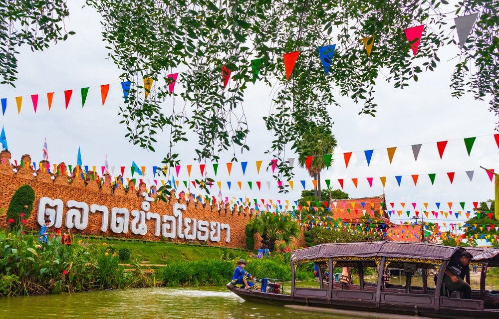 เรือลำหนึ่งลอยอยู่บนคลองหน้าตลาดนัดกลางแจ้งที่เต็มไปด้วยบรรยากาศครึกครื้น มีธงสีสันสดใสและป้ายภาษาไทยขนาดใหญ่บนผนังอิฐ สะท้อนให้เห็นถึงบรรยากาศที่มีชีวิตชีวาของ ตลาดน้ำใกล้กรุงเทพ