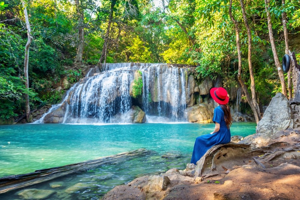 ผู้หญิง ที่เที่ยวธรรมชาติ ในชุดเดรสสีน้ำเงินและหมวกสีแดงนั่งอยู่บนก้อนหินข้างสระน้ำสีฟ้าคราม หันหน้าไปทางน้ำตกที่ล้อมรอบด้วยต้นไม้สีเขียว—เหมาะสำหรับผู้ที่กำลังมองหาสถานที่ท่องเที่ยวทางธรรมชาติ.