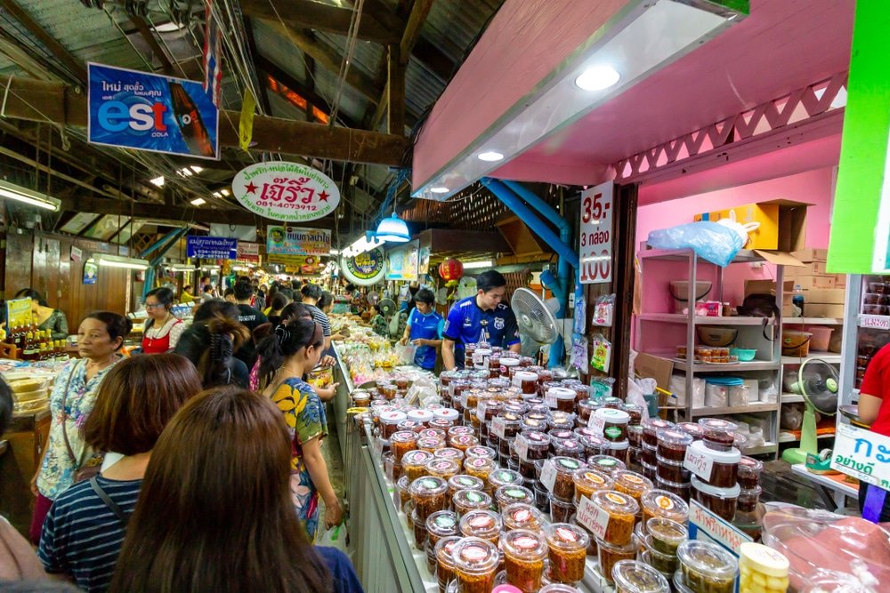 ผู้ซื้อของเดินชมและซื้อสินค้าจากผู้ขายในตลาดในร่มที่คึกคัก ซึ่งชวนให้นึกถึง ตลาดน้ำใกล้กรุงเทพ โดยมีขวดโหลของสินค้าที่เก็บรักษาไว้จัดแสดงอยู่บนเคาน์เตอร์.