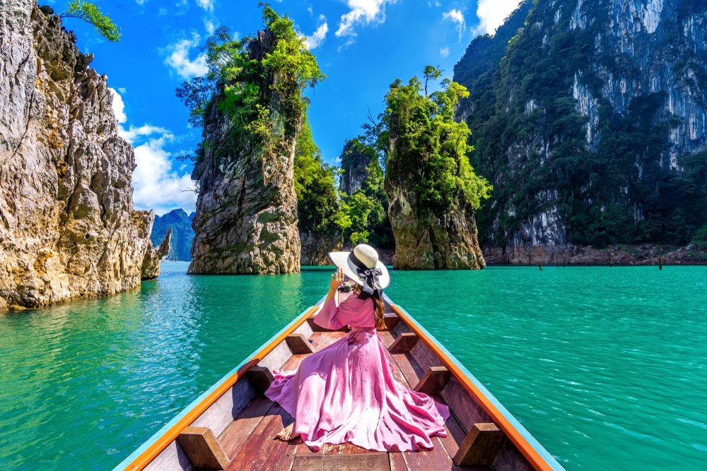 บุคคลในชุดเดรสและหมวกสีชมพู นั่งอยู่ด้านหน้าของเรือไม้ในน้ำสีฟ้าคราม กำลังสำรวจสถานที่ท่องเที่ยว ที่เที่ยวธรรมชาติ ทางธรรมชาติที่ล้อมรอบด้วยหน้าผาหินปูนสูงใหญ่และเขียวชอุ่ม ภายใต้ท้องฟ้าสีคราม.