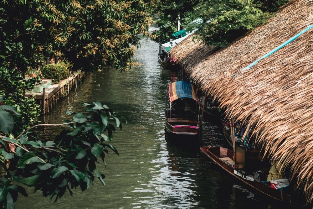เรือที่มีหลังคาสีสันสดใสจอดเรียงรายอยู่ตามคลองที่ขนาบข้างด้วยต้นไม้และอาคารหลังคามุงจากในช่วงเวลากลางวันที่ ตลาดน้ำใกล้กรุงเทพ มอบภาพชีวิตท้องถิ่นที่มีสีสันสดใส.
