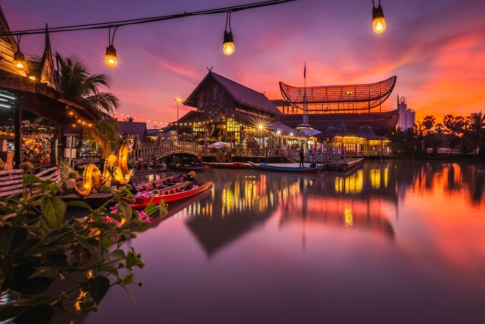 ตลาดริมแม่น้ำยามพระอาทิตย์ตกดิน ท้องฟ้าสีสันสดใส อาคารไม้แบบดั้งเดิม เรือลำน้อยลอยอยู่บนผืนน้ำ และแสงไฟประดับระยิบระยับเหนือศีรษะ—สัมผัสเสน่ห์ของ ตลาดน้ำใกล้กรุงเทพ