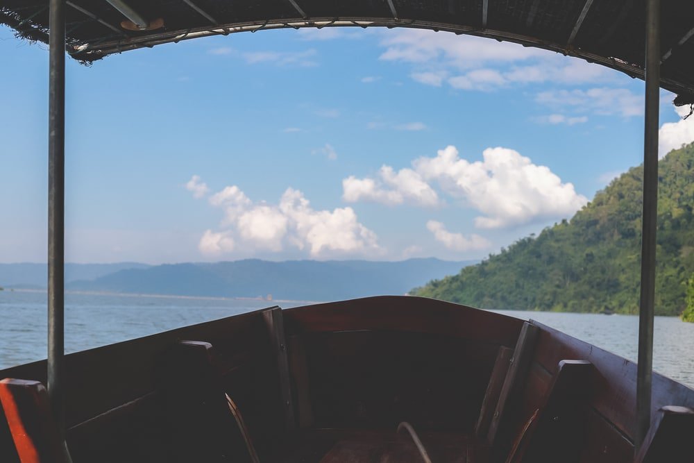 มุมมองจากภายในเรือไม้ที่มองออกไปยังทะเลสาบซึ่งมีภูเขาไกลๆ และ ที่เที่ยวธรรมชาติ ท้องฟ้าที่มีเมฆบางส่วนเป็นฉากหลัง โดยมีหลังคาเรือเป็นกรอบภาพ—เหมาะสำหรับผู้ที่มองหาสถานที่ท่องเที่ยวทางธรรมชาติและความสงบเหนือผืนน้ำ.
