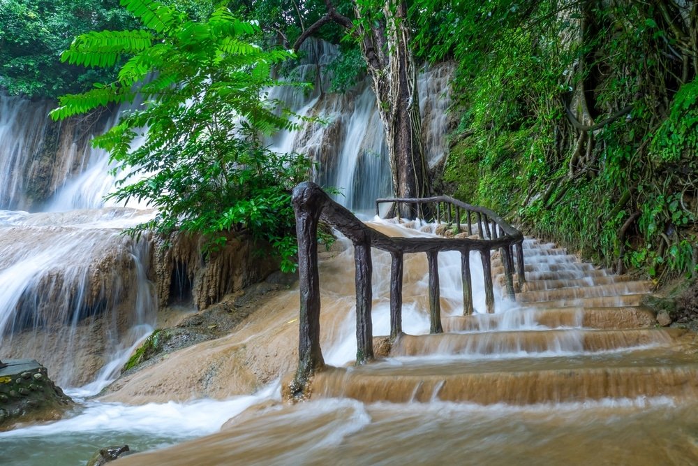 บันไดหินพร้อมราวจับไม้ทอดยาวขึ้นข้างน้ำตกที่ไหลลดหลั่นลงมา ล้อมรอบด้วยพืชพรรณเขียวชอุ่มใน ที่เที่ยวธรรมชาติ บรรยากาศป่าไม้—เหมาะสำหรับผู้ที่กำลังมองหาสถานที่ท่องเที่ยวทางธรรมชาติ.