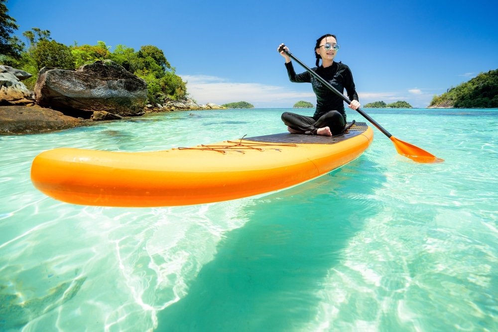 บุคคลในชุดสีดำและแว่นกันแดดกำลังพายเรือคายัคสีส้มบนผืนน้ำสีฟ้าใสใกล้ ที่เที่ยวธรรมชาติ ชายฝั่งหินใต้ท้องฟ้าแจ่มใส เพลิดเพลินกับหนึ่งในสถานที่ท่องเที่ยวธรรมชาติยอดนิยม.