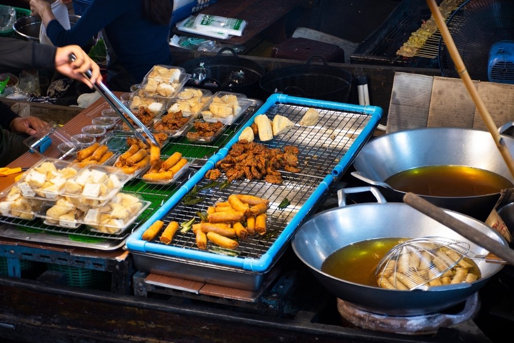 พ่อค้าแม่ค้าขายอาหารริมทางที่ ตลาดน้ำใกล้กรุงเทพ กำลังเตรียมและจัดแสดงของว่างทอดหลากหลายชนิดบนตะแกรงลวดข้างกระทะใบใหญ่สองใบที่เต็มไปด้วยน้ำมัน ณ แผงขายของที่คึกคักในตลาด.