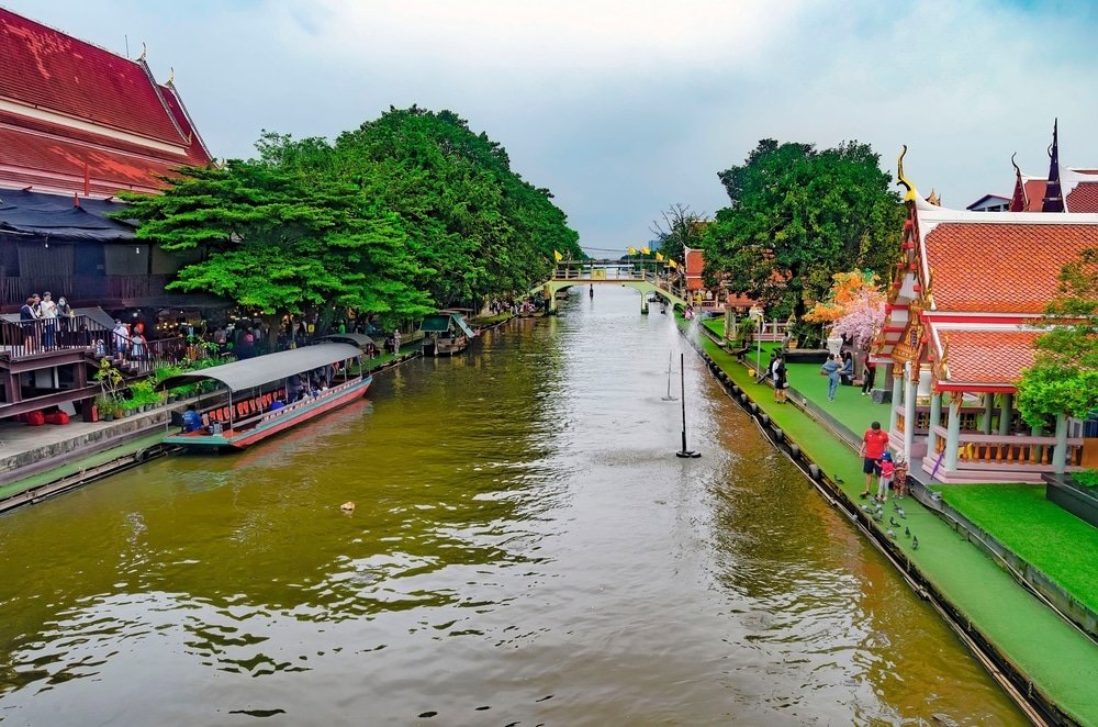 คลองที่ขนาบข้างด้วยต้นไม้และอาคารแบบดั้งเดิม มีเรือยาวลอยอยู่บนน้ำและผู้คนเดินไปตามทางเดิน—ถ่ายทอดบรรยากาศที่มีชีวิตชีวาของ ตลาดน้ำใกล้กรุงเทพ