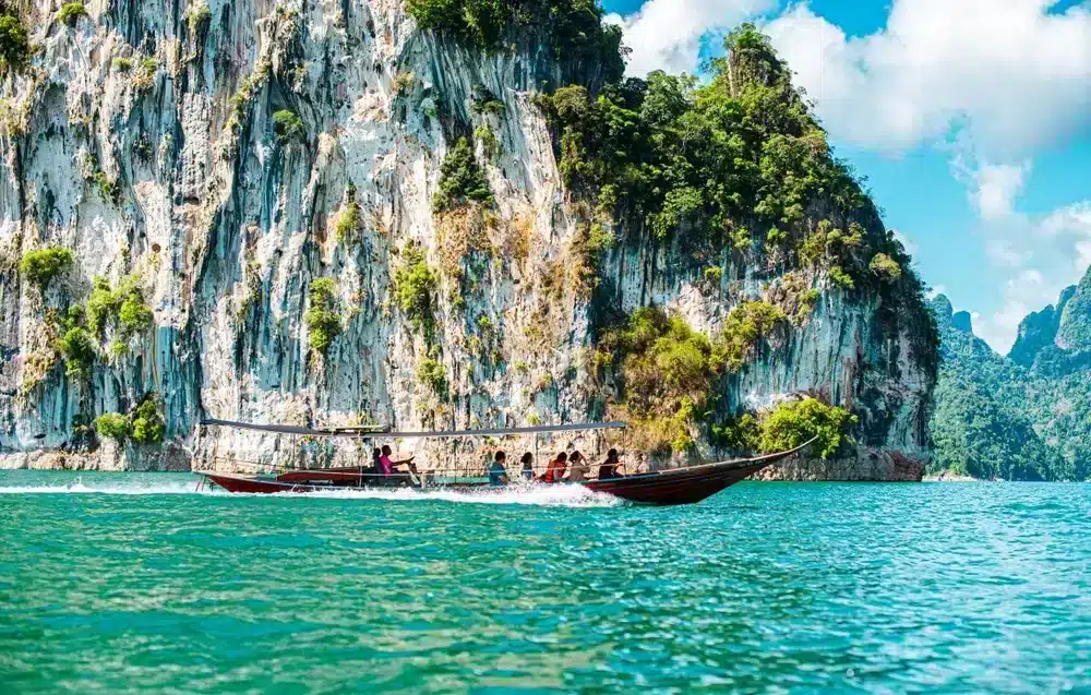 เรือหางยาวที่มีผู้โดยสารหลายคนแล่นผ่านน้ำสีฟ้าครามเบื้องหน้า ที่เที่ยวธรรมชาติ หน้าผาหินปูนสูงชันที่ปกคลุมไปด้วยต้นไม้ ท่ามกลางท้องฟ้าที่มีเมฆบางส่วน—เป็นสถานที่ท่องเที่ยวทางธรรมชาติที่น่าทึ่ง เหมาะสำหรับผู้ที่ชื่นชอบการผจญภัย.