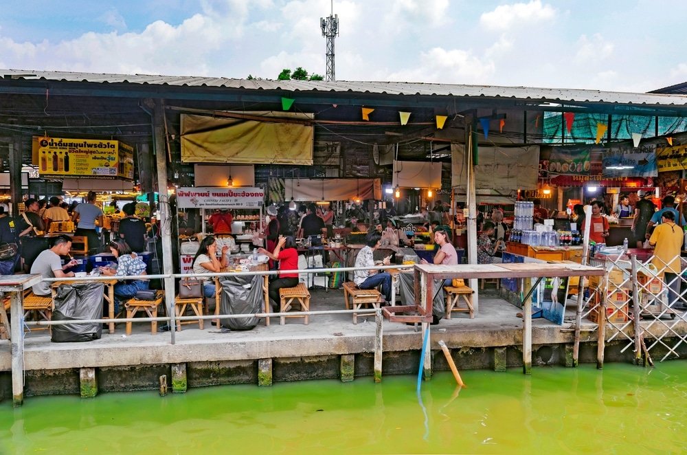 ตลาดอาหารกลางแจ้งที่มีผู้คนรับประทานอาหารที่โต๊ะริมทางเดินริมน้ำ มีร้านค้าและป้ายสีสันสดใสภายใต้พื้นที่ที่มีหลังคาคลุม—เหมาะสำหรับผู้ที่ต้องการบรรยากาศ ตลาดน้ำใกล้กรุงเทพ ที่คึกคัก.