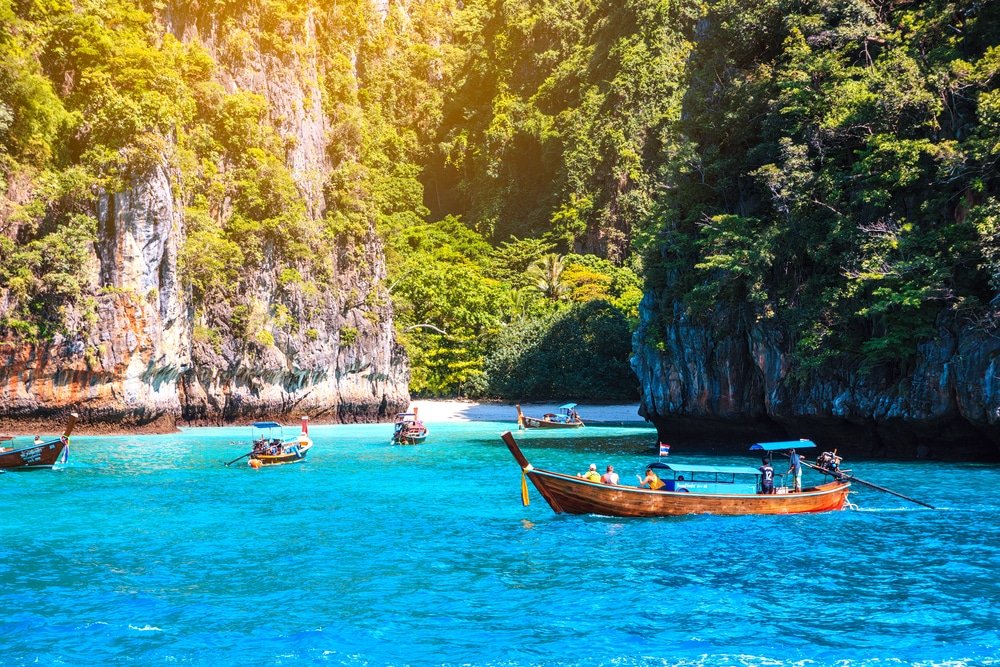 เรือไม้ลอยอยู่บนน้ำทะเลใสสีเทอร์ควอยซ์ใกล้ชายหาดทรายเล็ก ๆ ล้อมรอบด้วยหน้าผาสูงใหญ่สีเขียวสดใสภายใต้แสงแดดจ้า—เหมาะสำหรับผู้ ที่เที่ยวธรรมชาติ ที่ชื่นชอบการท่องเที่ยวธรรมชาติ.