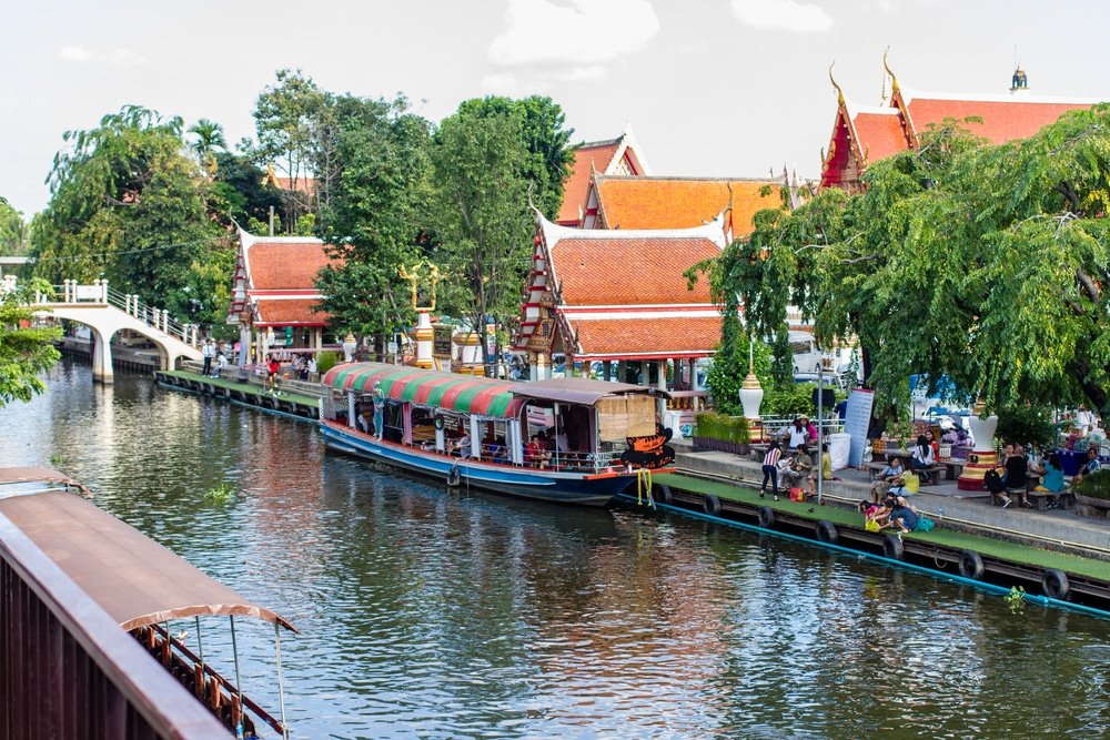 เรือหลากสีสันจอดเรียงรายอยู่ริมคลองข้างอาคารไทยดั้งเดิมที่มีหลังคาสีแดง ต้นไม้ และผู้คนกำลังเพลิดเพลินกับบรรยากาศที่มีชีวิตชีวาของ ตลาดน้ำใกล้กรุงเทพ ริมแม่น้ำ.