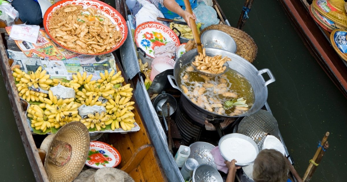 พ่อค้าแม่ค้าบนเรือที่ ตลาดน้ำใกล้กรุงเทพ (ตลาดน้ำใกล้กรุงเทพ) ทอดและขายอาหาร รวมถึงกล้วยและของว่างชุบแป้งทอด โดยใช้หม้อโลหะขนาดใหญ่บนเตา.
