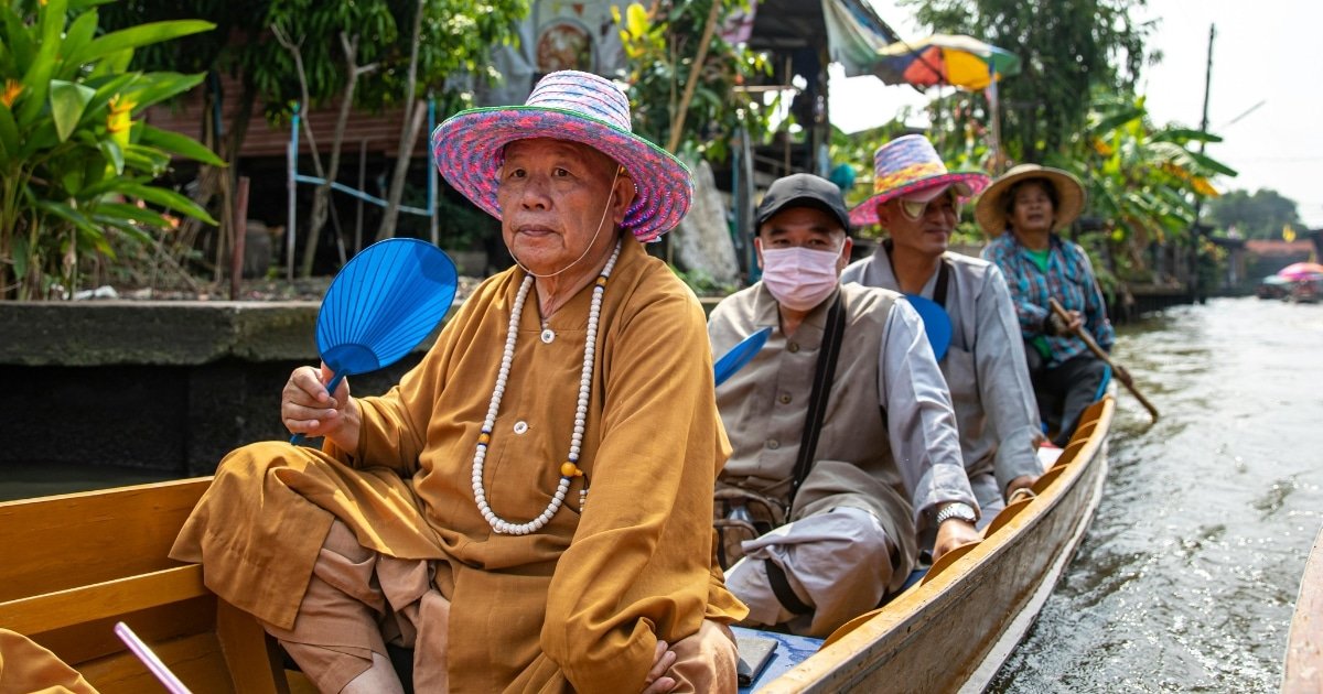 กลุ่มคนสวมหมวกนั่งอยู่ในเรือไม้ล่องไปตามคลองที่ ตลาดน้ำใกล้กรุงเทพ ล้อมรอบด้วยต้นไม้และอาคาร.