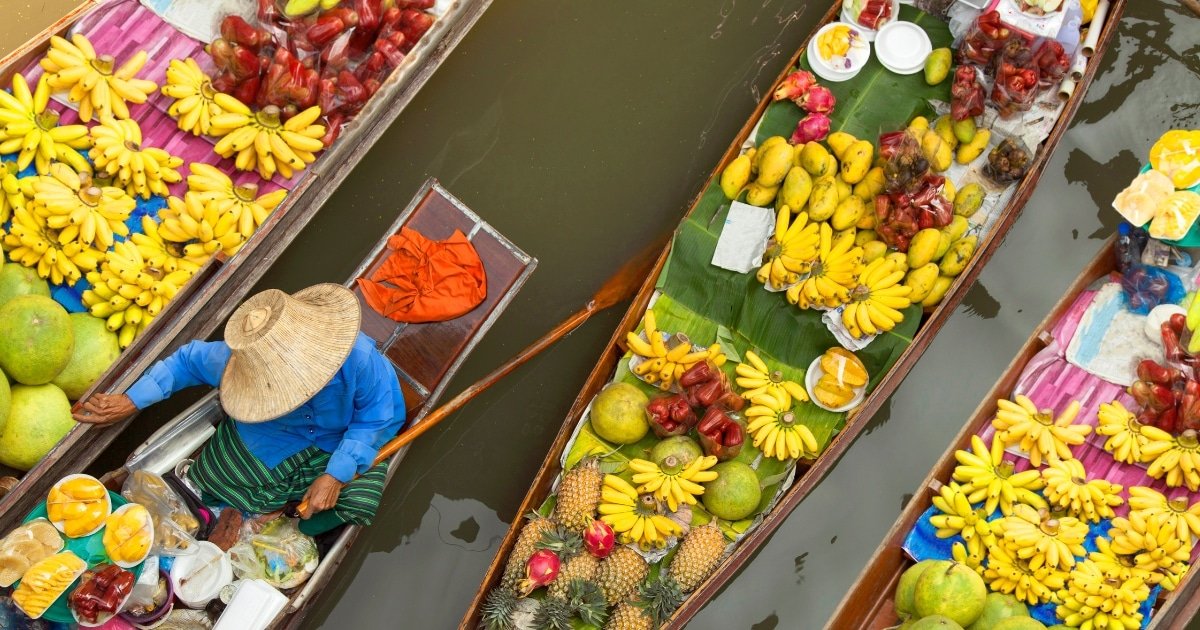 ภาพมุมสูงของเรือไม้ที่ ตลาดน้ำใกล้กรุงเทพ เต็มไปด้วยกล้วย มะม่วง สับปะรด และผลไม้ชนิดอื่น ๆ; มีพ่อค้าในเสื้อสีฟ้าและหมวกฟางนั่งอยู่ในเรือลำหนึ่ง.