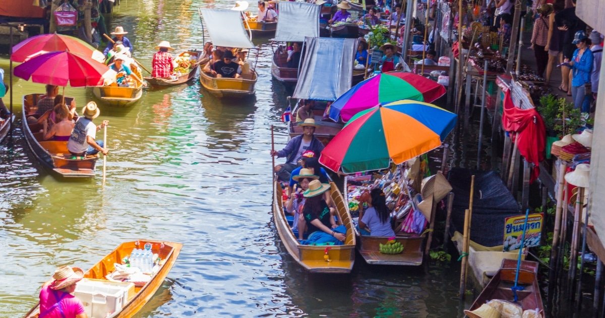 เรือที่มีร่มสีสันสดใสและพ่อค้าแม่ค้าขายสินค้าอยู่บนคลองตลาดน้ำที่คึกคัก ล้อมรอบด้วยผู้คนบนน้ำและริมฝั่งที่ ตลาดน้ำใกล้กรุงเทพ ที่เต็มไปด้วยชีวิตชีวา.