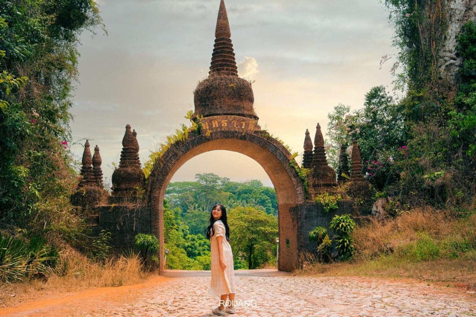 ที่เที่ยวสุราษฎร์ธานี หญิงสาวในชุดสีขาว ยืนอยู่บนทางเดินที่ปูด้วยก้อนหินกลม หน้าซุ้มประตูหินโบราณ รายล้อมไปด้วยต้นไม้ใหญ่และพืชพรรณเขียวชอุ่ม สร้างบรรยากาศเสน่ห์ของสถานที่ท่องเที่ยวสุราษฎร์ธานี.