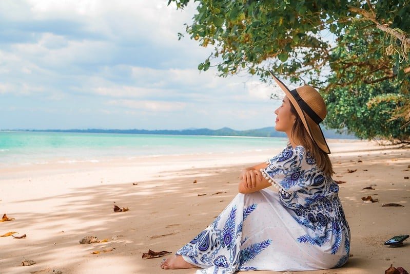 ผู้หญิงคนหนึ่งสวมหมวกกันแดดและชุดลาย นั่งอยู่บนชายหาดทรายใต้ต้นไม้ ณ สถานที่ท่องเที่ยวทางธรรมชาติ มองออกไปยังทะเลที่มีเมฆและแผ่นดินไกลๆ เป็นฉากหลัง.