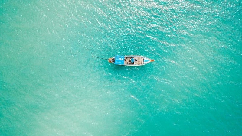 ภาพมุมสูงของเรือลำเดียวลอยอยู่บนน้ำสีฟ้าใสราวกับเทอร์ควอยซ์ เหมาะสำหรับผู้ที่มองหาสถานที่ท่องเที่ยวทางธรรมชาติและต้องการสัมผัสความสงบกลางทะเล.