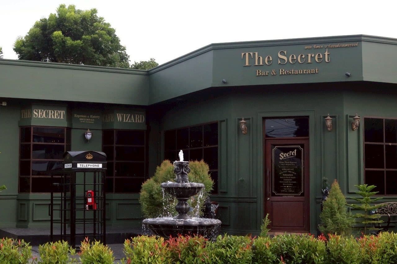 อาคารสีเขียวในสุราษฎร์ธานีที่เที่ยว มี "The Secret Bar & Restaurant" อยู่ภายใน พร้อมน้ำพุสีดำอยู่ด้าน ที่เที่ยวสุราษฎร์ธานี หน้า ตู้โทรศัพท์อยู่ทางซ้าย และต้นไม้เขียวชอุ่มล้อมรอบทางเข้า.