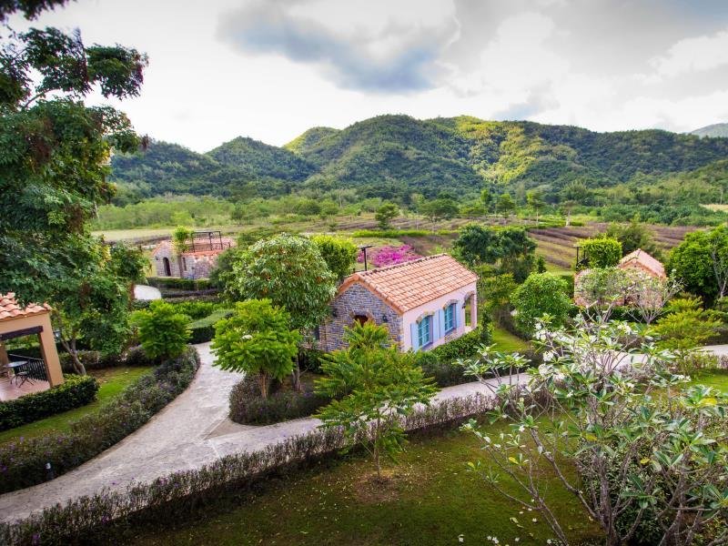 บ้านหลังเล็กหลังหนึ่งที่มีหลังคามุงกระเบื้องสีแดงและบานหน้าต่าง ที่เที่ยวปลายฝนต้นหนาว สีน้ำเงินล้อมรอบด้วยต้นไม้และทางเดิน—สถานที่ท่องเที่ยวที่น่าดึงดูดในช่วงปลายฤดูฝนต้นฤดูหนาวพร้อมภูเขาเขียวขจีและท้องฟ้าที่มีเมฆลอยเป็นฉากหลัง.