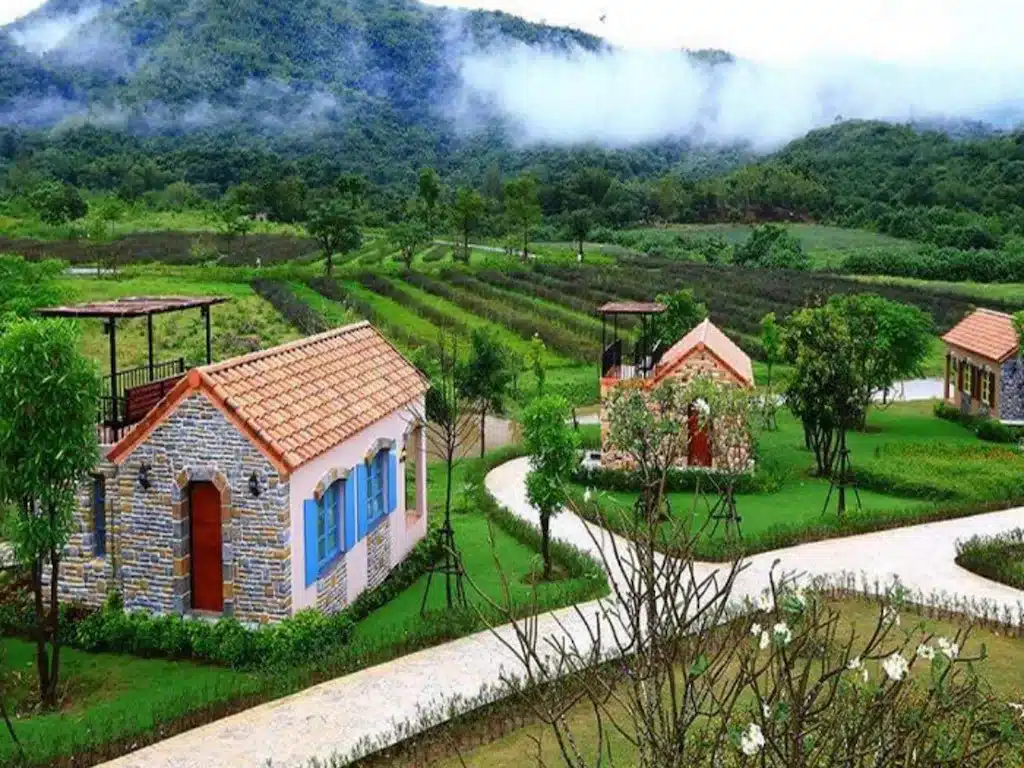 กระท่อมหินเล็ก ๆ หลังคาสีแดงตั้งอยู่ท่ามกลางสนามหญ้าสีเขียวและ ที่เที่ยวปลายฝนต้นหนาว ทางเดินคดเคี้ยว ล้อมรอบด้วยเนินเขาและหมอกในทิวทัศน์ชนบท—เหมาะสำหรับคนรักธรรมชาติและการท่องเที่ยวปลายฝนต้นหนาว.