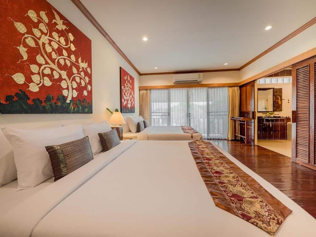 ห้องพักโรงแรมกว้างขวางพร้อมเตียงใหญ่สองเตียง, งานศิลปะ ที่เที่ยวปลายฝนต้นหนาว ตกแต่งสีแดงและทอง, พื้นไม้, และมุมนั่งเล่นสบาย ๆ ใกล้หน้าต่างพร้อมผ้าม่านบางเบา—เหมาะสำหรับการผ่อนคลายหลังจากสำรวจสถานที่ท่องเที่ยวปลายฝนต้นหนาว.