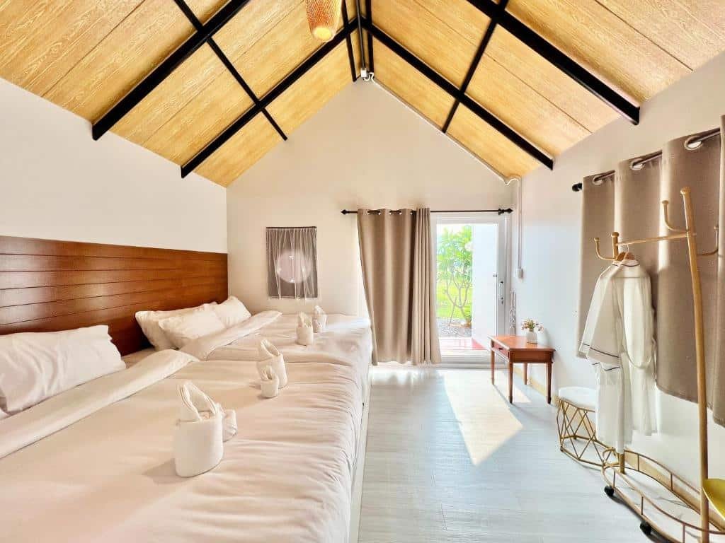 ห้องนอนสว่างไสวพร้อมเตียงใหญ่สองเตียง ผ้าเช็ดตัวพับไว้เพดานไม้ ประตูเปิดโล่งสู่ภายนอก และราว ที่เที่ยวปลายฝนต้นหนาว แขวนเสื้อพร้อมเสื้อคลุม—เหมาะสำหรับการผ่อนคลายหลังจากท่องเที่ยวตามสถานที่ท่องเที่ยวปลายฝนต้นหนาว แสงแดดส่องผ่านเข้ามาทางประตูและหน้าต่าง.