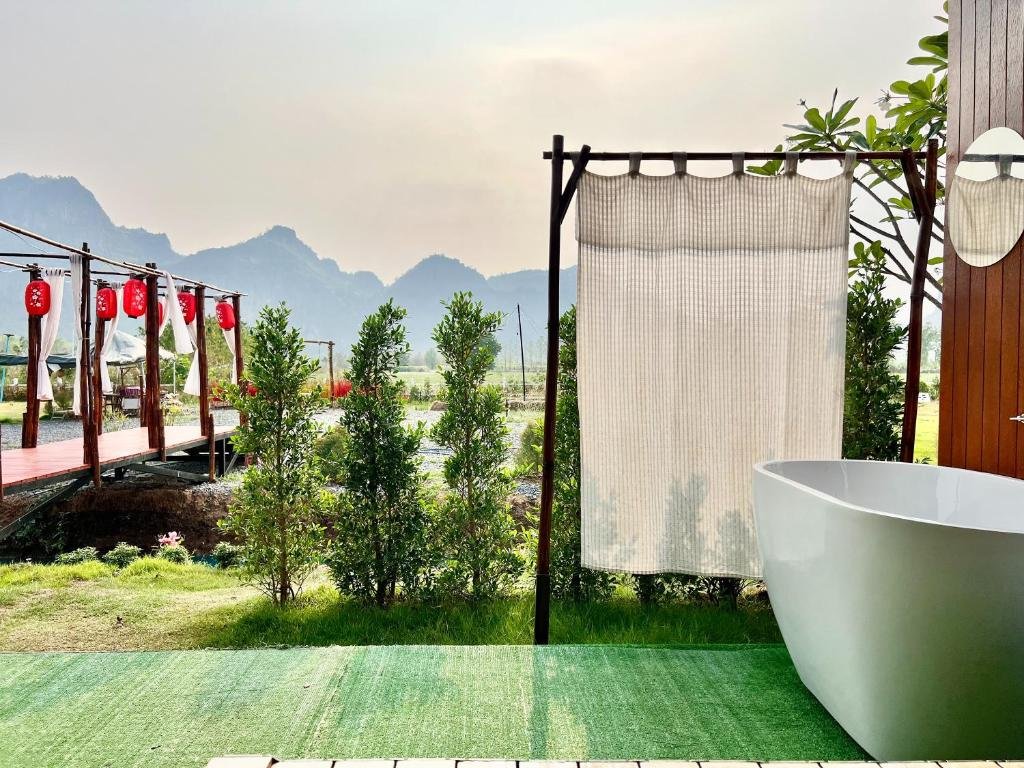 อ่างอาบน้ำกลางแจ้งตั้งอยู่ริมม่านบังตาเพื่อความเป็นส่วนตัว มองเห็นทิวทัศน์สีเขียวขจี ทางเดินไม้ ที่เที่ยวปลายฝนต้นหนาว ประดับโคมแดง และภูเขาไกลสุดสายตา—สถานที่พักผ่อนในฤดูฝนปลายปีสู่ต้นหนาวที่สมบูรณ์แบบ ท่ามกลางท้องฟ้าหมอกบาง.