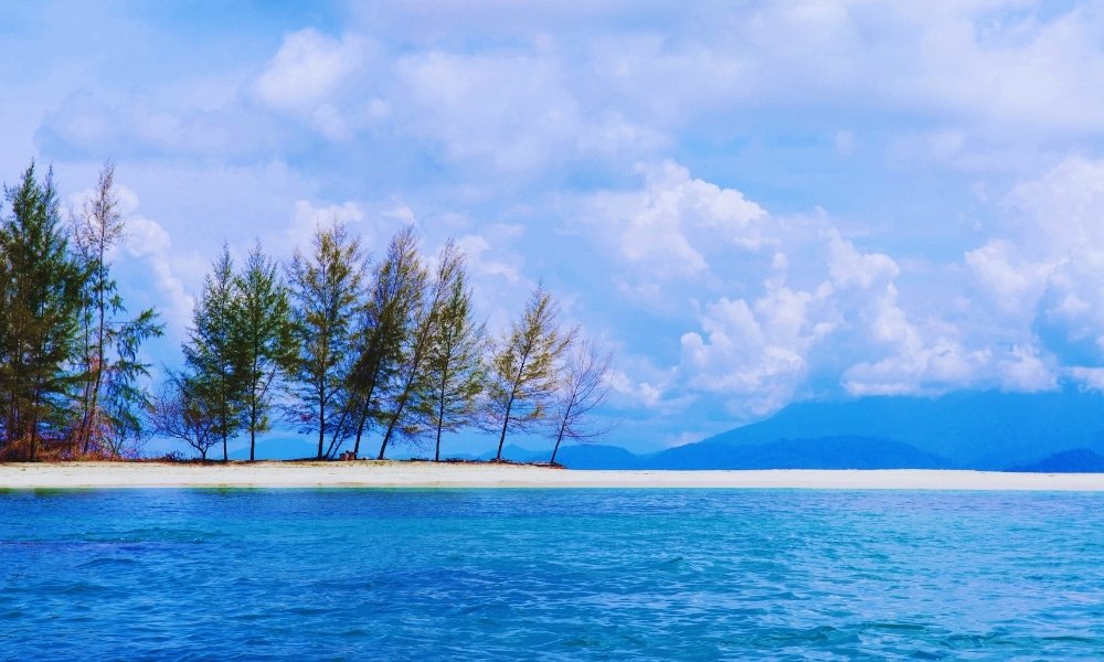 เกาะทรายเล็ก ๆ ที่มีต้นสนเรียงรายอยู่ริมชายหาด ล้อมรอบด้วยน้ำทะเลใสสีฟ้าภายใต้ท้องฟ้าที่มีเมฆบางส่วน ทำให้เป็นสถานที่ท่องเที่ยวทางธรรมชาติที่เหมาะสำหรับผู้ที่ต้องการความสงบและความสวยงามตามธรรมชาติ.