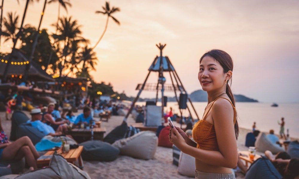 ผู้หญิงคนหนึ่งยืนอยู่บนชายหาดที่มีชีวิตชีวาในยามพระอาทิตย์ตกดินที่ ที่เที่ยวสุราษฎร์ธานี สุราษฎร์ธานีที่เที่ยว รายล้อมไปด้วยผู้คนนั่งพักผ่อนบนถุงฟองน้ำและที่นั่งกลางแจ้งใกล้ทะเล.
