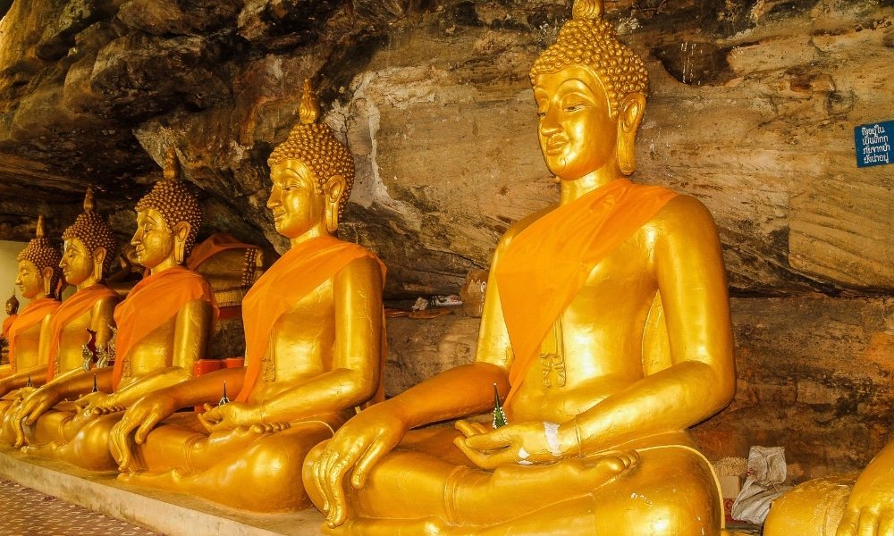พระพุทธรูปทองคำเรียงรายประดับด้วยผ้าสีส้ม นั่งในท่าสมาธิพิงผนัง เที่ยวสุราษฎร์ธานี ถ้ำหิน—ภาพอันเงียบสงบที่ สุราษฎร์ธานีที่เที่ยว เหมาะสำหรับผู้ที่ต้องการสำรวจสถานที่ศักดิ์สิทธิ์ขณะเที่ยวสุราษฎร์ธานี.