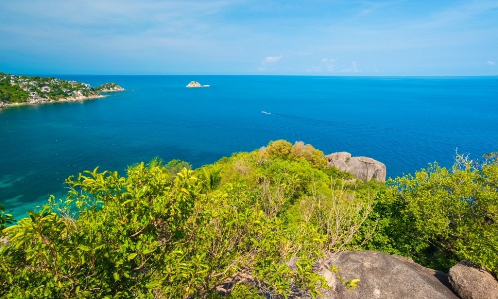 เพลิดเพลินกับวิวทะเลสีฟ้าใส เที่ยวสุราษฎร์ธานี สวยงาม เรือลำเล็ก ชายฝั่งหิน และพืชพรรณเขียวชอุ่ม พร้อมเกาะที่อยู่ไกลออกไป—เหมาะสำหรับผู้ที่มองหาสถานที่ท่องเที่ยวในสุราษฎร์ธานีภายใต้ท้องฟ้าที่สดใส.