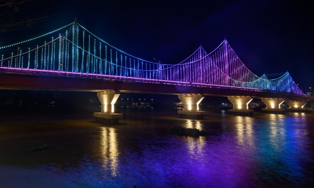 สะพานแขวนที่ประดับด้วยไฟ LED สีสันสดใสทอดข้ามแม่น้ำในยาม เที่ยวสุราษฎร์ธานี ค่ำคืน สร้างภาพสะท้อนที่งดงาม—สถานที่ท่องเที่ยวที่ไม่ควรพลาดในสุราษฎร์ธานีสำหรับผู้ที่ต้องการสำรวจความงามของเมืองนี้หลังพระอาทิตย์ตกดิน.