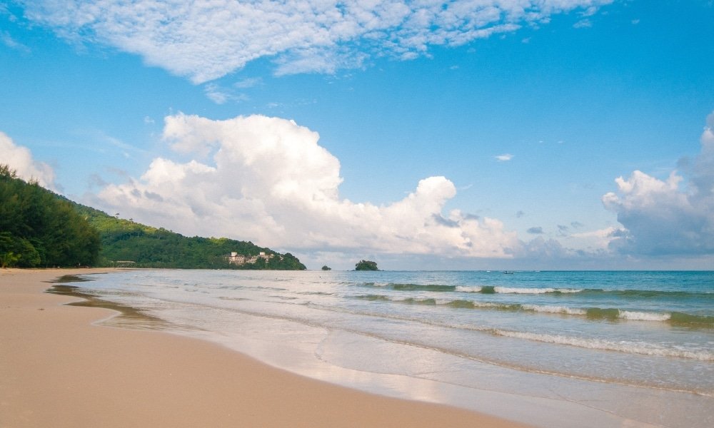 ชายหาดทรายที่มีคลื่นเบาและทะเลสงบ ภูเขาเขียวขจีอยู่ทางซ้าย และท้องฟ้าสีฟ้าที่มีเมฆบางส่วน—เหมาะสำหรับผู้ที่มองหา สถานที่ท่องเที่ยวสุราษฎร์ธานี หรือสำรวจสถานที่ท่องเที่ยวสุราษฎร์ธานี.