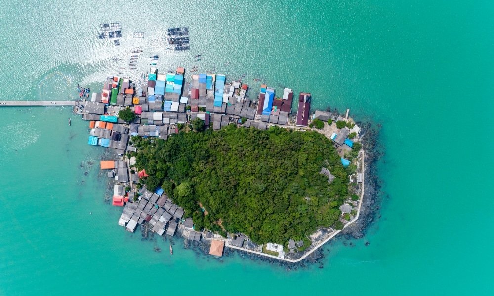 ภาพมุมสูงของเกาะเล็ก ๆ ที่มีพืช ที่เที่ยวสุราษฎร์ธานี พรรณเขียวชอุ่มหนาแน่นอยู่ตรงกลาง และอาคารหลากสีสันเรียงรายตามแนวชายฝั่ง ล้อมรอบด้วยน้ำทะเลสีฟ้าเทอร์ควอยซ์—เหมาะสำหรับผู้ที่กำลังมองหากิจกรรมท่องเที่ยวสุราษฎร์ธานีและประสบการณ์ใหม่ ๆ ในสุราษฎร์ธานี.