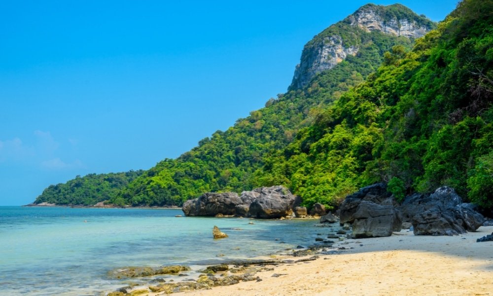 ชายหาดทรายขาวสะอาด น้ำทะเลใสสีฟ้า แนวชายฝั่งที่เต็มไปด้วยโขดหิน และเนินเขาเขียวขจีภายใต้ ที่เที่ยวสุราษฎร์ธานี ท้องฟ้าแจ่มใส—เหมาะสำหรับผู้ที่กำลังค้นหาสถานที่ท่องเที่ยวในสุราษฎร์ธานีและฝันถึงการหลบหนีจากความวุ่นวายเพื่อพักผ่อนริมทะเล.
