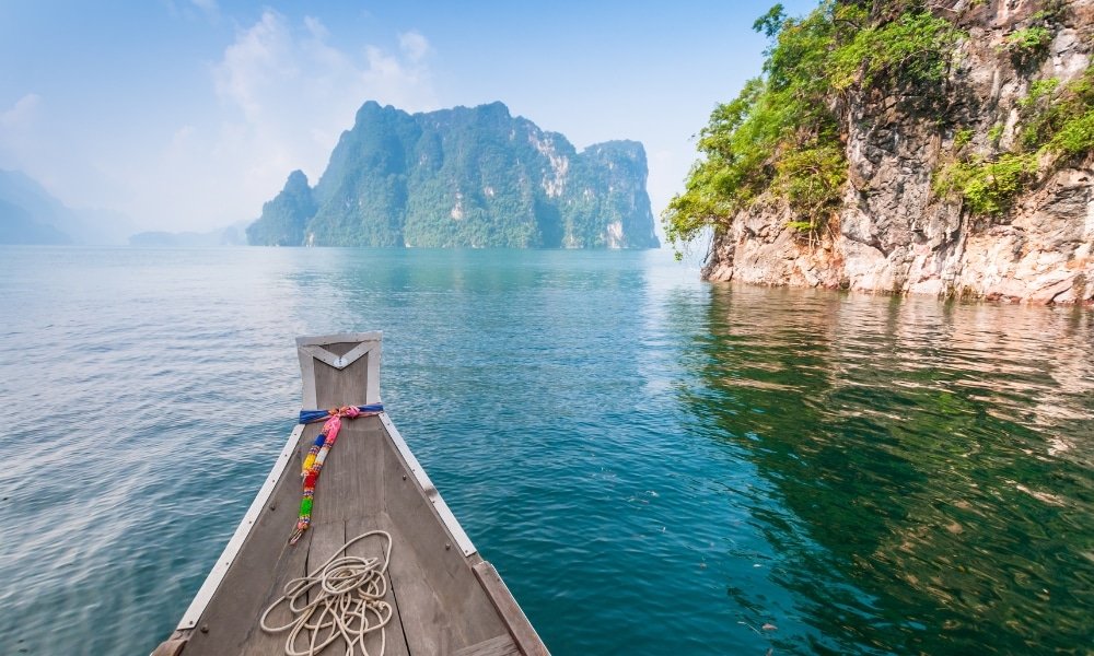 เรือไม้ลำหนึ่งมีเชือกผูกอยู่ที่หัวเรือ ลอยอยู่บนผืนน้ำสีฟ้าใส ท่ามกลางหน้าผาหินและต้นไม้เขียวขจี—ภาพทิวทัศน์อันเงียบสงบ เหมาะอย่างยิ่งสำหรับผู้ที่กำลังค้นหา สถานที่ท่องเที่ยวในสุราษฎร์ธานี ท่ามกลางขุนเขาสีเขียวไกลสุดสายตาและท้องฟ้าที่มีเมฆบางส่วน.