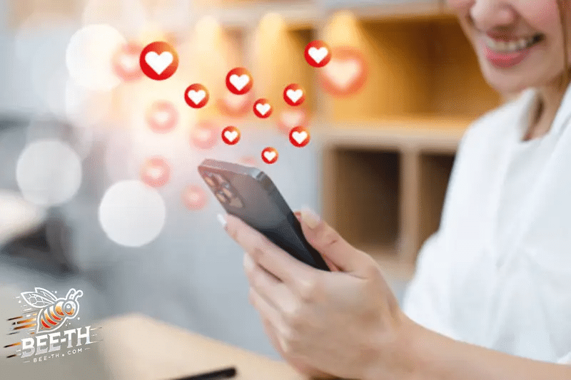 ผู้หญิงคนหนึ่งยิ้มขณะใช้สมาร์ทโฟนของเธอ โดยมีไอคอนรูปหัวใจลอยอยู่รอบๆ ซึ่งแสดงถึงการกดไลค์หรือการตอบสนอง—ถ่ายทอดผลลัพธ์ของเว็บปั๊มวิวได้อย่างสมบูรณ์แบบ โลโก้ของ Beeth.com ปรากฏอยู่ที่มุมล่างซ้าย.