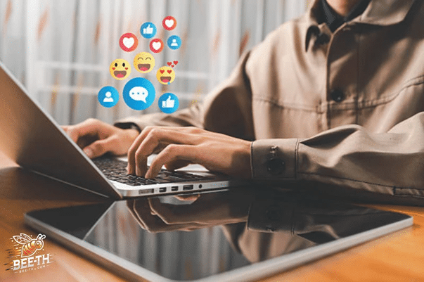 บุคคลกำลังพิมพ์บนแล็ปท็อป โดยมีไอคอนปฏิกิริยาจากโซเชียลมีเดียลอยอยู่เหนือหน้าจอ แสดงให้เห็นถึงการมีส่วนร่วมที่อาจมาจาก เว็บปั้มวิว; แท็บเล็ตวางอยู่บนโต๊ะข้างๆ.