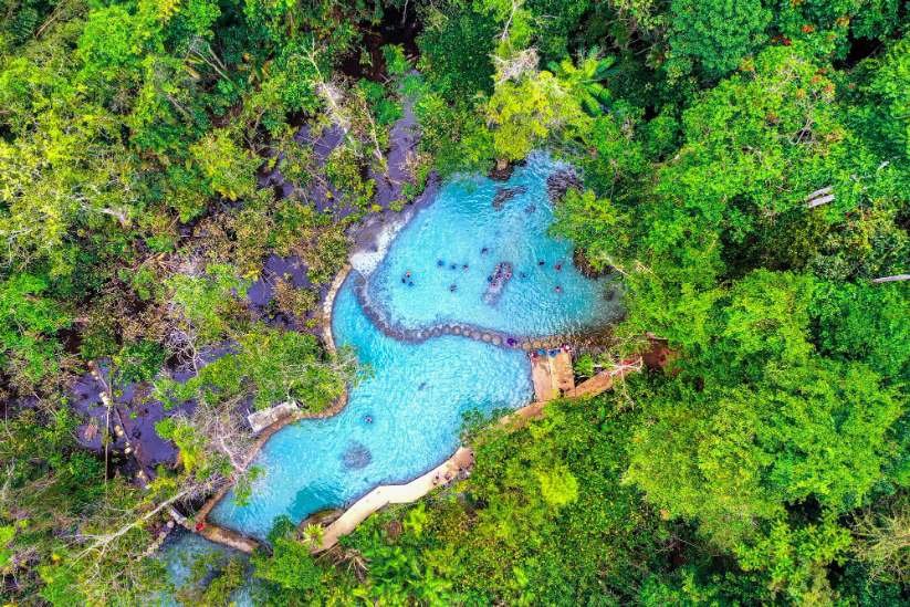ภาพมุมสูงของสระน้ำสีฟ้าธรรมชาติที่ล้อมรอบด้วยป่าเขียวชอุ่มหนาแน่น มีผู้คนกำลังว่ายน้ำและทางเดินไม้ทอดยาวตามขอบสระ—สถานที่ท่องเที่ยวทางธรรมชาติที่สมบูรณ์แบบสำหรับการพักผ่อนและการผจญภัย.