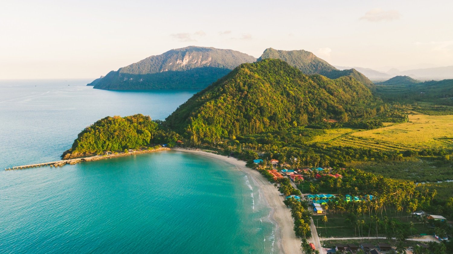 ภาพมุมสูงของชายหาดทรายโค้งที่สวยงาม เป็นแหล่งท่องเที่ยวทางธรรมชาติที่งดงามล้อมรอบด้วยน้ำทะเลสีฟ้าคราม ภูเขาเขียวขจี และชุมชนชายฝั่งขนาดเล็กที่มีอาคารสีสันสดใส.