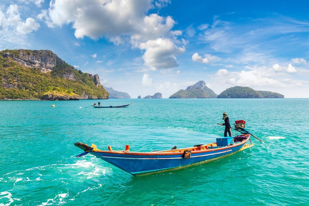 บุคคลหนึ่งยืนอยู่บนเรือไม้ในน้ำสีฟ้าคราม ณ สถานที่ท่องเที่ยว ที่เที่ยวสุราษฎร์ธานี สุราษฎร์ธานี โดยมีเกาะหินและท้องฟ้าสีฟ้าที่มีเมฆเป็นฉากหลัง เรืออีกลำหนึ่งปรากฏอยู่ไกลออกไป เน้นเสน่ห์ของการท่องเที่ยวสุราษฎร์ธานี.