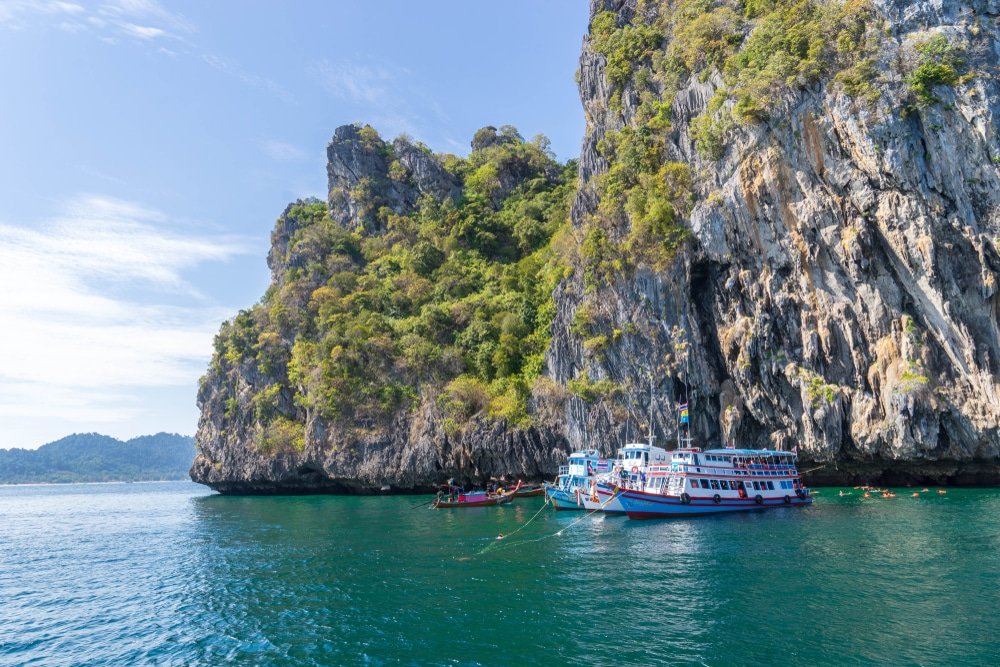 เกาะหินขนาดใหญ่ที่มีพืชพรรณสีเขียวขจีโผล่ขึ้นมาจากทะเล มีเรือหลากสีจอดอยู่ใกล้ฐานหน้าผาบนผืนน้ำสีฟ้าสงบ ทำให้ที่นี่กลายเป็นสถานที่ท่องเที่ยวทางธรรมชาติที่สวยงามตระการตาสำหรับนักท่องเที่ยวที่แสวงหาความงดงามของธรรมชาติ.