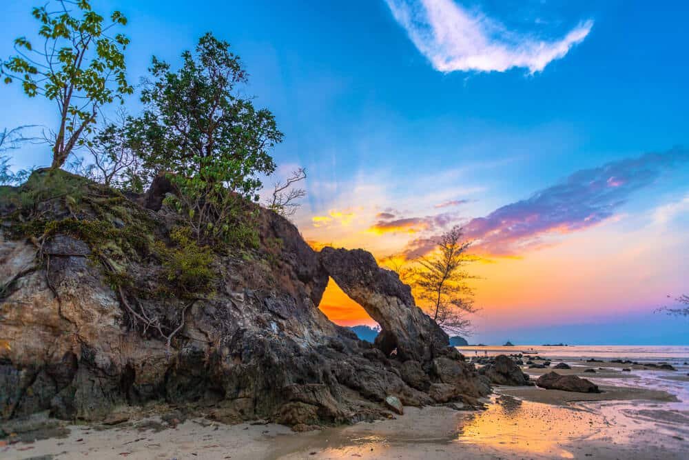 หินรูปร่างสวยงามที่มีซุ้มประตูธรรมชาติตั้งอยู่บนชายหาดทราย ล้อมรอบด้วยต้นไม้และแอ่งน้ำตามธรรมชาติ ท่ามกลางท้องฟ้าที่มีเมฆบางส่วนในยามพระอาทิตย์ขึ้นหรือพระอาทิตย์ตก—เหมาะสำหรับผู้ที่ชื่นชอบการท่องเที่ยวธรรมชาติและต้องการความงามอันเงียบสงบ.