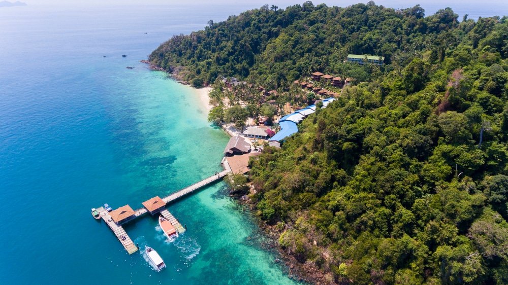 ภาพมุมสูงของรีสอร์ทบนเกาะเขตร้อน หนึ่งในสถานที่ท่องเที่ยวทางธรรมชาติยอดนิยม มีเรือจอดอยู่ที่ท่าเรือไม้ ล้อมรอบด้วยน้ำทะเลสีฟ้าใสและป่าเขียวชอุ่ม.